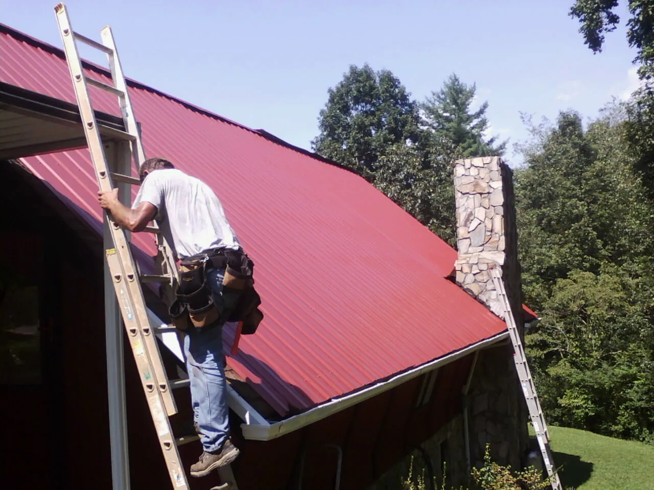 Roofer on a ladder beside a red metal roof and stone chimney — Soffit & Fascia Repair by Roofing Brothers in Yellville